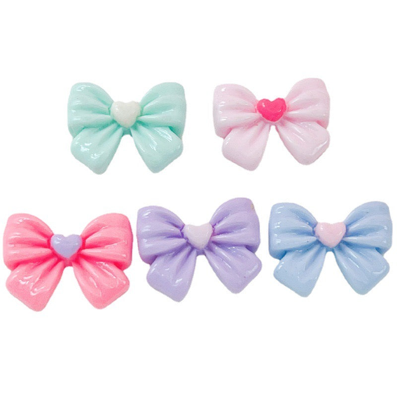 These are pastel-colored bow cabochons in shades of mint green, light pink, bright pink, lavender, and sky blue. Each bow features a small, heart-shaped embellishment at the center, adding a cute detail to the design. These cabochons might be used for a variety of crafting purposes, such as decorating phone cases, jewelry making, scrapbooking, or embellishing other small items.
