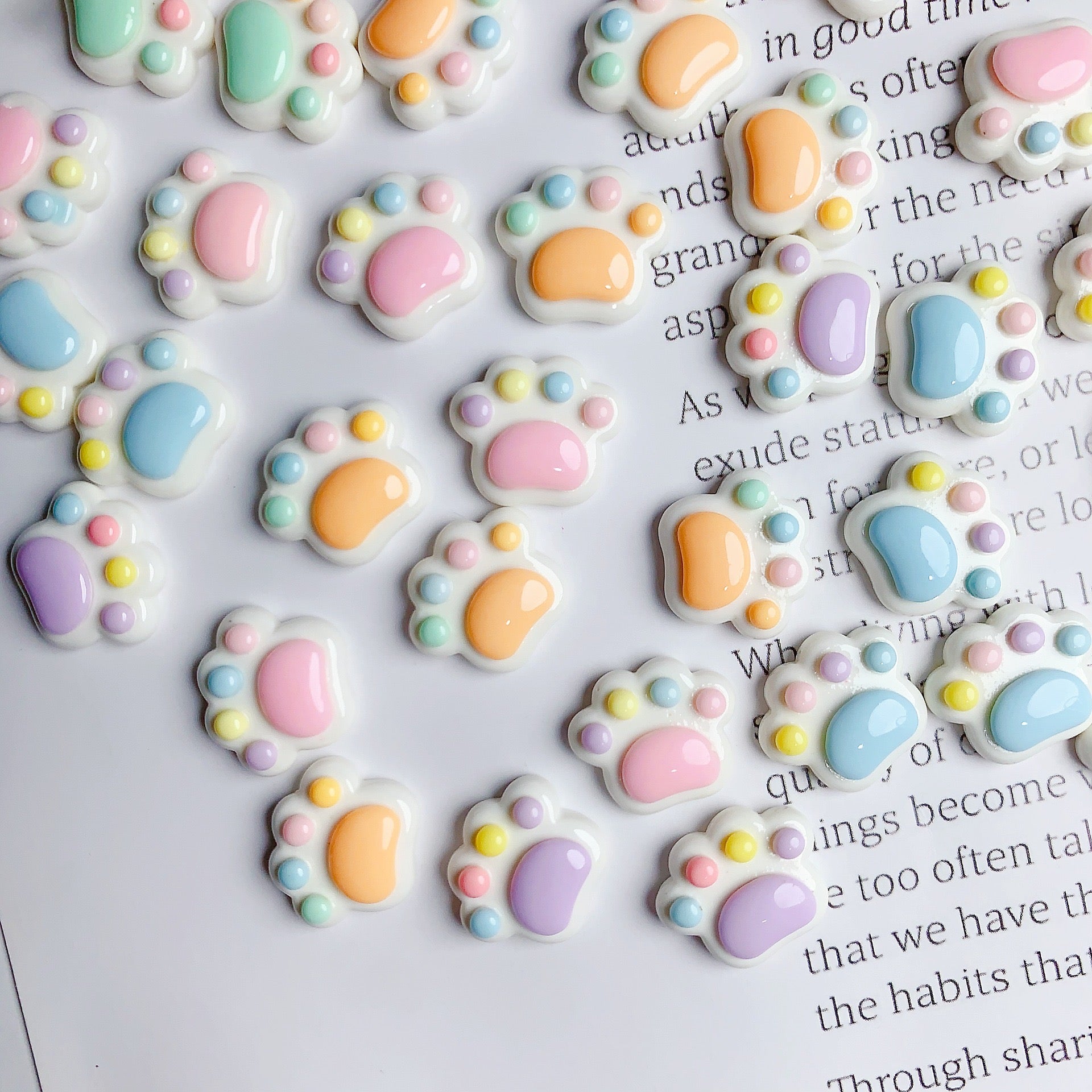 These are colorful paw print cabochons with a whimsical and cute design. They feature a variety of pastel colors for the paw pads and are adorned with small, bead-like accents in complementary colors around the larger pads. The cabochons are set on a white background that appears to be a page with text, suggesting they might be used for crafting or as decorations on top of a paper surface. These could be ideal for creating unique accessories, decorating scrapbooks, customizing personal items, or for use in 